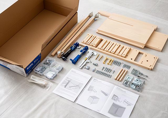 Furniture assembly parts and instruction sheets arranged on a protected floor beside an opened flat-pack carton